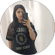 Small circular profile photo of a woman taking a mirror selfie with a smartphone, wearing a dark printed T‑shirt and standing against a light background, used as a casual avatar or reviewer image.