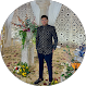 Small circular profile photo of a man in a patterned suit standing in a decorated venue with flowers and a stylish backdrop, used as a formal avatar or reviewer image.